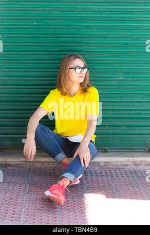 Beautiful woman wears t-shirt, blue jeans and sunglasses sitting on the pedestrian area near city building. She looks tired Stock Photo