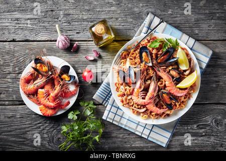 Spanish Fideua, a noodle Paella with king prawns, white fish meat, calamari, mussels served on a white platter on a wooden table with roasted seafoods Stock Photo