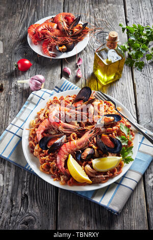 Spanish Fideua, a noodle Paella with king prawns, white fish meat, calamari, mussels served on a white platter on a wooden table with roasted seafoods Stock Photo