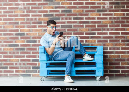 Full length portrait of shocked young bearded man in casual style and eyeglasses sitting on blue wooden pallet, holding smartphone and reading unbelie Stock Photo