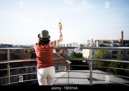 The young pretty brunette stands on the balcony and holds a cool drink in her hands. Stock Photo