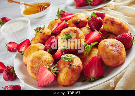 cottage cheese balls served with strawberries on a white plate on a wooden table with honey, sour cream and fresh strawberries in a bowl at the backgr Stock Photo