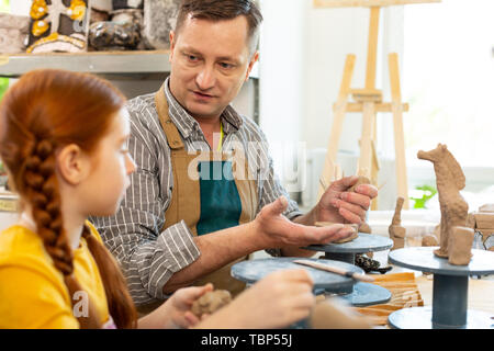 Teacher wearing apron. Art teacher wearing apron talking to his little pupil while working with clay Stock Photo