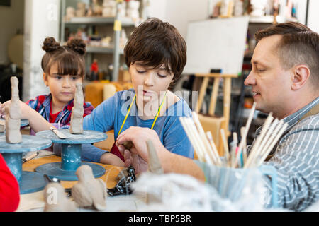 Listening to teacher. Dark-haired brother and sister listening to their art teacher while sculpting clay animals Stock Photo