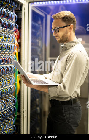Serious concentrated qualified bearded programmer in glasses standing by server cabinet and using laptop while examining server Stock Photo