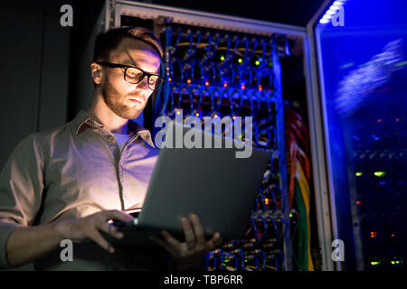 Serious busy young IT engineer in glasses standing against network server equipment with glowing sensors in dark room and using laptop while monitorin Stock Photo