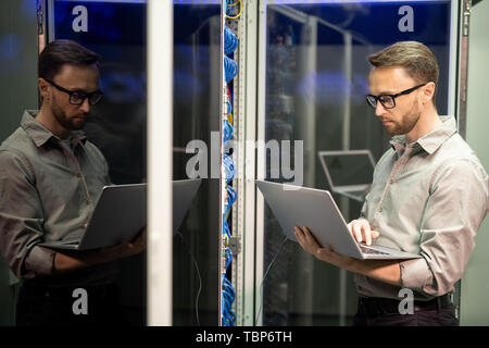 Serious busy young IT engineer with beard standing by server cabinet and using laptop while providing network support Stock Photo