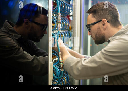 Side view of concentrated young server repairman in glasses standing in server room and connecting cables to supercomputer Stock Photo