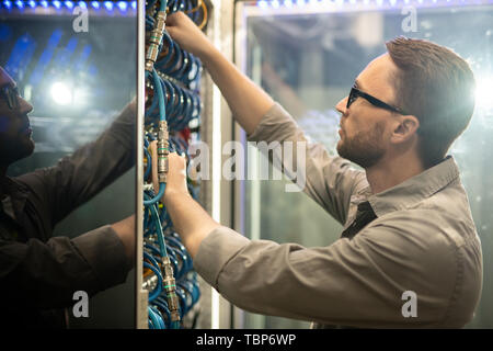 Serious skilled young data center engineer in glasses standing at mainframe and examining cables while doing checkup Stock Photo
