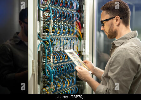 Serious pensive young data center technician in glasses standing against cabinet of server and using digital tablet while checking connections Stock Photo
