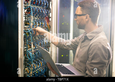 Serious busy young male engineer in glasses standing against mainframe and adjusting cables while analyzing server connections Stock Photo