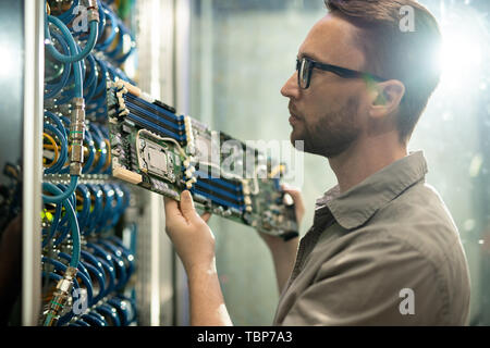 Serious skilled young bearded server installation specialist in glasses holding circuit board and examining server racks while working in datacenter r Stock Photo
