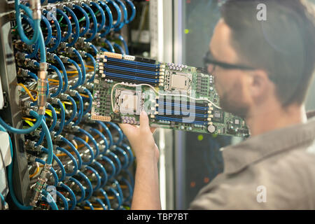Busy skilled young IT expert in glasses standing by server cabinet and analyzing motherboard of supercomputer Stock Photo
