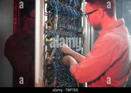Serious busy young IT specialist in glasses standing by powerful mainframe and examining cables while maintaining networking system Stock Photo