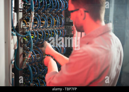 Rear view of skilled man standing by cabinet of server racks and examining fiber cables while repairing server Stock Photo