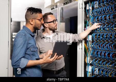 Serious thoughtful young multi-ethnic computer support specialists in casual clothing standing at server racks and using laptop while analyzing networ Stock Photo