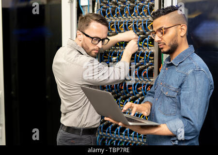 Serious young multi-ethnic IT engineers in casual shirts standing in database center and setting up computer cluster while examining information on la Stock Photo