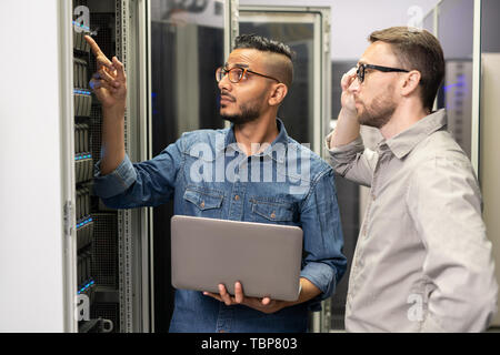 Serious thoughtful young multi-ethnic network engineers in glasses and casual shirts standing at cabinet and working with supercomputer Stock Photo