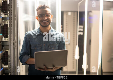Portrait of cheerful satisfied young middle-eastern server engineer in glasses standing in datacenter room and using laptop Stock Photo