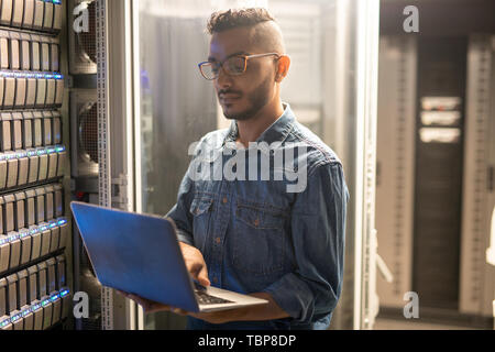 Serious concentrated young middle-eastern server specialist in glasses standing at network server and using portable computer while doing network moni Stock Photo