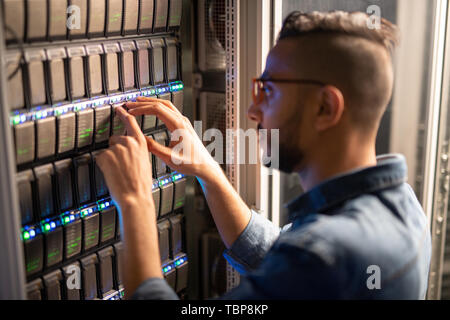 Serious busy hipster IT specialist standing by open cabinet and examining network racks while analyzing provider of server Stock Photo