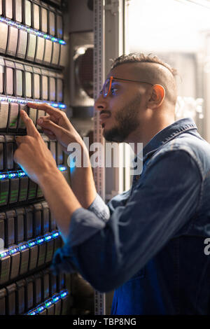 Serious pensive young middle-eastern server support engineer in eyeglasses standing at modern server and analyzing sensors of server equipment Stock Photo