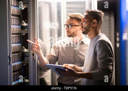Team of young multi-ethnic specialists in glasses standing at server and using tablet while managing network server together in data center Stock Photo
