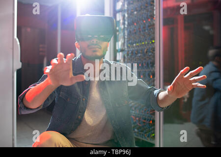 Serious concentrated young Arabian IT engineer with beard crouching on floor of data center and gesturing hands while studying server systems using VR Stock Photo