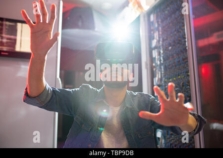Serious modern IT specialist with beard working in data center and monitoring server systems while using virtual reality goggles, sunlight effect Stock Photo