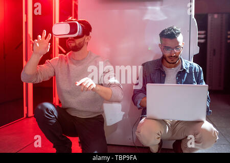 Serious curious young multi-ethnic IT engineers sitting on floor in data center and testing new app for augmented reality Stock Photo