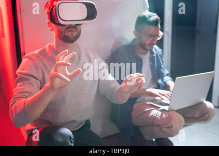 Arabian IT engineer typing on laptop while transferring data to colleague in VR goggles in data center Stock Photo
