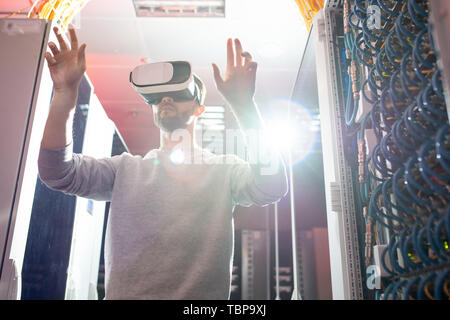 Serious concentrated young IT scientist in virtual reality goggles standing in server room and wirelessly interacting with rack servers Stock Photo