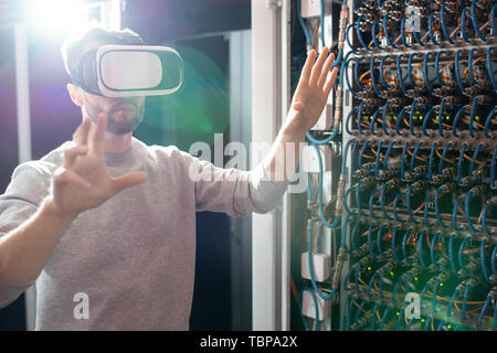 Serious concentrated young bearded engineer in VR goggles standing in database center and gesturing hands while managing server links wirelessly Stock Photo