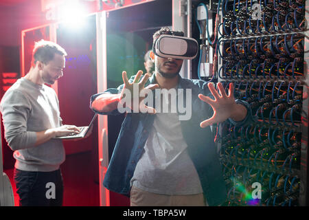 Concentrated hipster IT engineer in virtual reality goggles standing in server room and researching database server with augmented reality Stock Photo