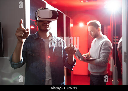 Serious hipster young middle-eastern IT scientist pointing with finger and using virtual reality simulator while testing server opportunities in data  Stock Photo