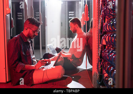Serious busy multi-ethnic IT engineers sitting on floor in server center and updating device configurations on laptops Stock Photo