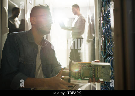 Serious young Arabian IT professional crouching in front of cabinet of mainframe and installing blade server, sunlight effect Stock Photo