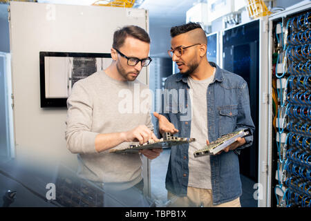 Serious thoughtful young multi-ethnic IT engineers in casual outfits standing in modern storage room and fixing motherboard together Stock Photo