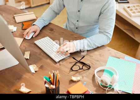 Close-up of unrecognizable creative web designer in casual shirt sitting at wooden table with messy crumpled papers and using desktop computer Stock Photo