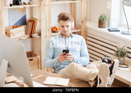Content relaxed young photographer in wired headphones sitting at table and keeping feet on table while using smartphone in design studio Stock Photo