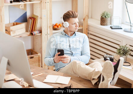 Serious pensive young guy in white headphones keeping feet on table and using smartphone while answering message and listening to music through headph Stock Photo