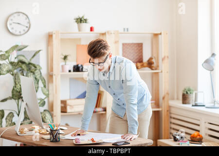 Serious pensive handsome young male engineer in glasses standing by table with modern computer and examining blueprint in office Stock Photo
