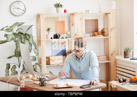 Serious skilled young male engineer in eyeglasses sitting at desk and using pair of compasses while measuring lines on blueprint Stock Photo