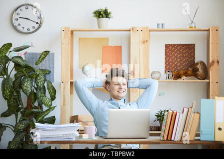 Happy inspired young office manager in light blue shirt sitting at table and keeping hands behind head while relaxing at workplace Stock Photo