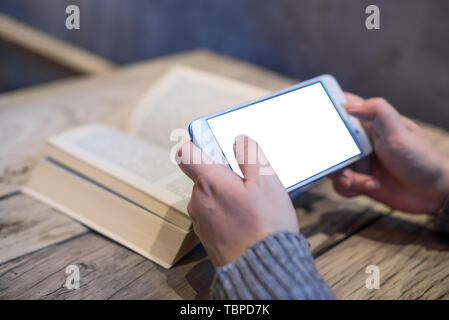 Woman holding a smartphone with an open book on a wooden table in a coffee shop Stock Photo