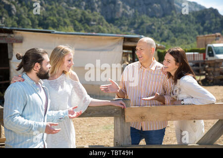 Happy adult friends breezily chatting near wooden fencing in country house in warm spring day Stock Photo