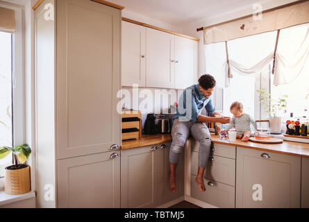 A father and a small toddler son eating fruit and yoghurt indoors at home. Stock Photo