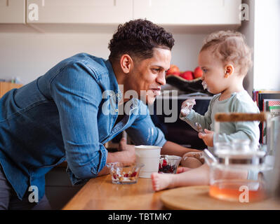 A father and a small toddler son eating fruit and yoghurt indoors at home. Stock Photo