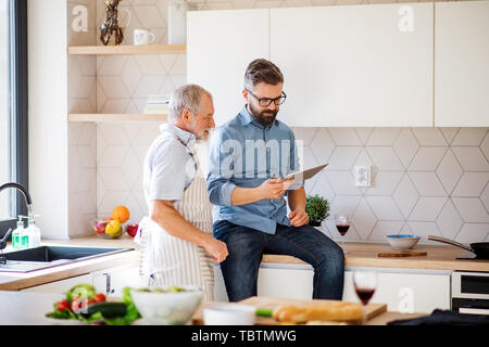 Adult hipster son and senior father indoors in kitchen at home, using tablet. Stock Photo