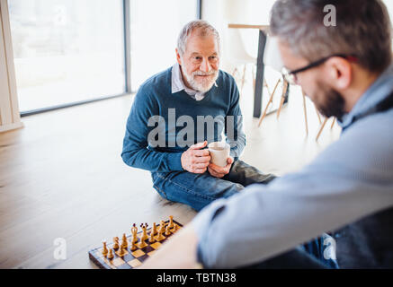 An adult hipster son and senior father sitting on floor indoors at home, playing chess. Stock Photo